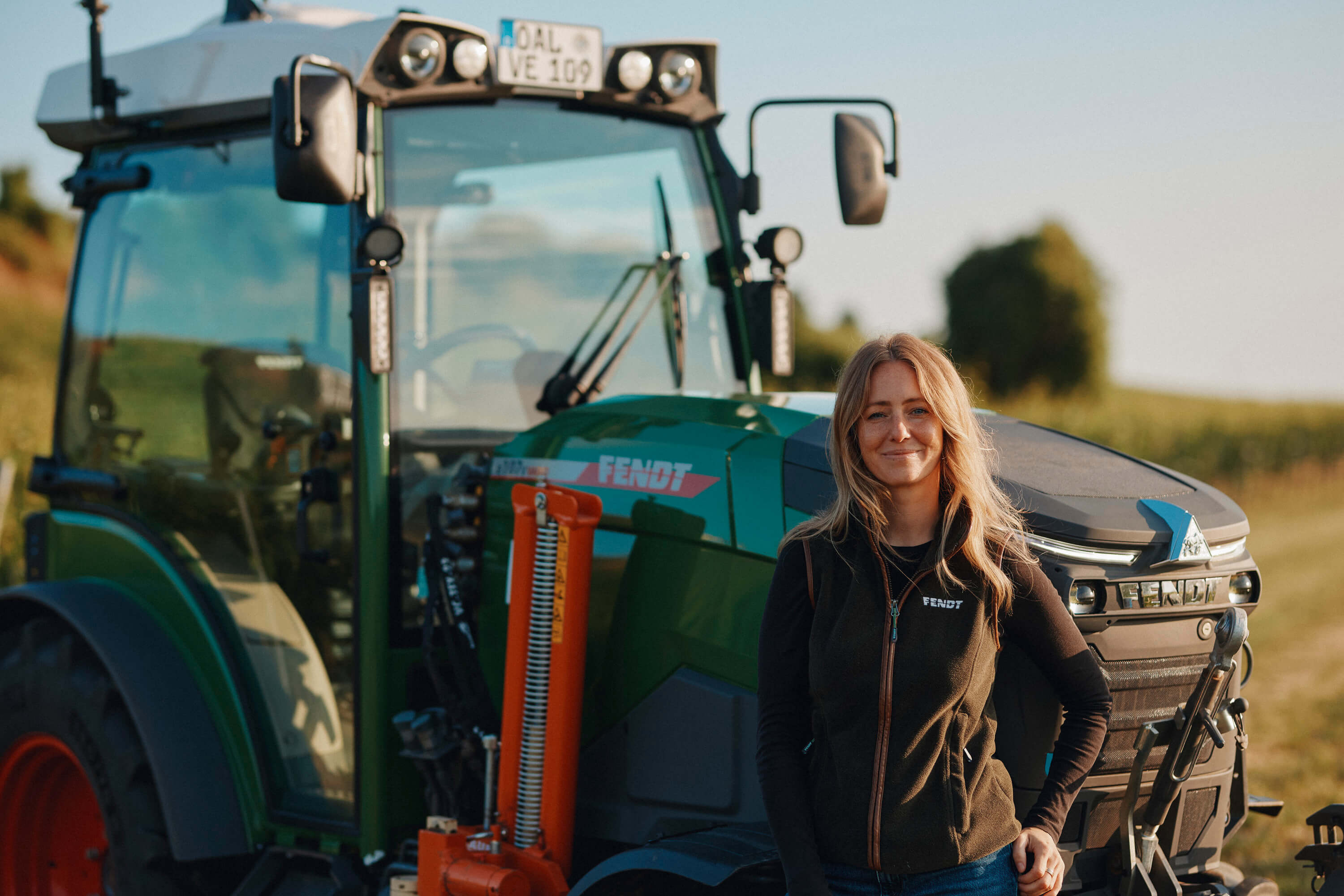 Juliane Eller, Gründerin von JUWEL Weine, steht vor dem gründen Fendt e107 V Vario Traktor mit roten Felgen. Sie trägt eine dunkle Fendt Weste, im Hintergrund sieht man die Weinberge.
