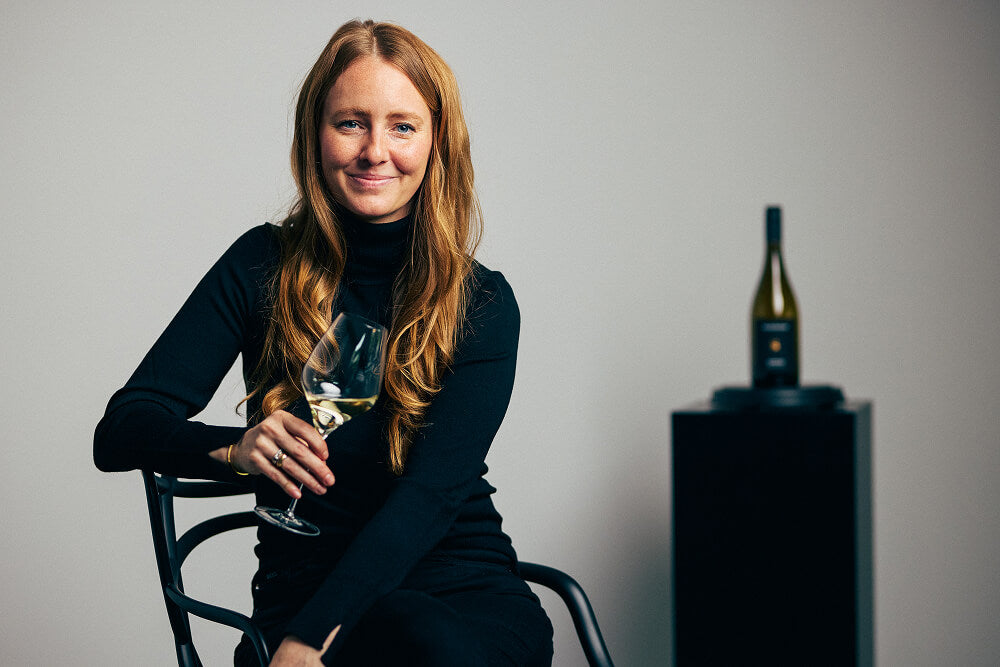 Juliane Eller, Gründerin von JUWEL Weine, im schwarzen BOSS Outfit auf einem Stuhl sitzend. Sie hält ein Glas Weißwein in der Hand, im Hintergrund ist eine Flasche des JULIANE ELLER PRESENTED BY BOSS | GRAUBURGUNDER zu sehen.
