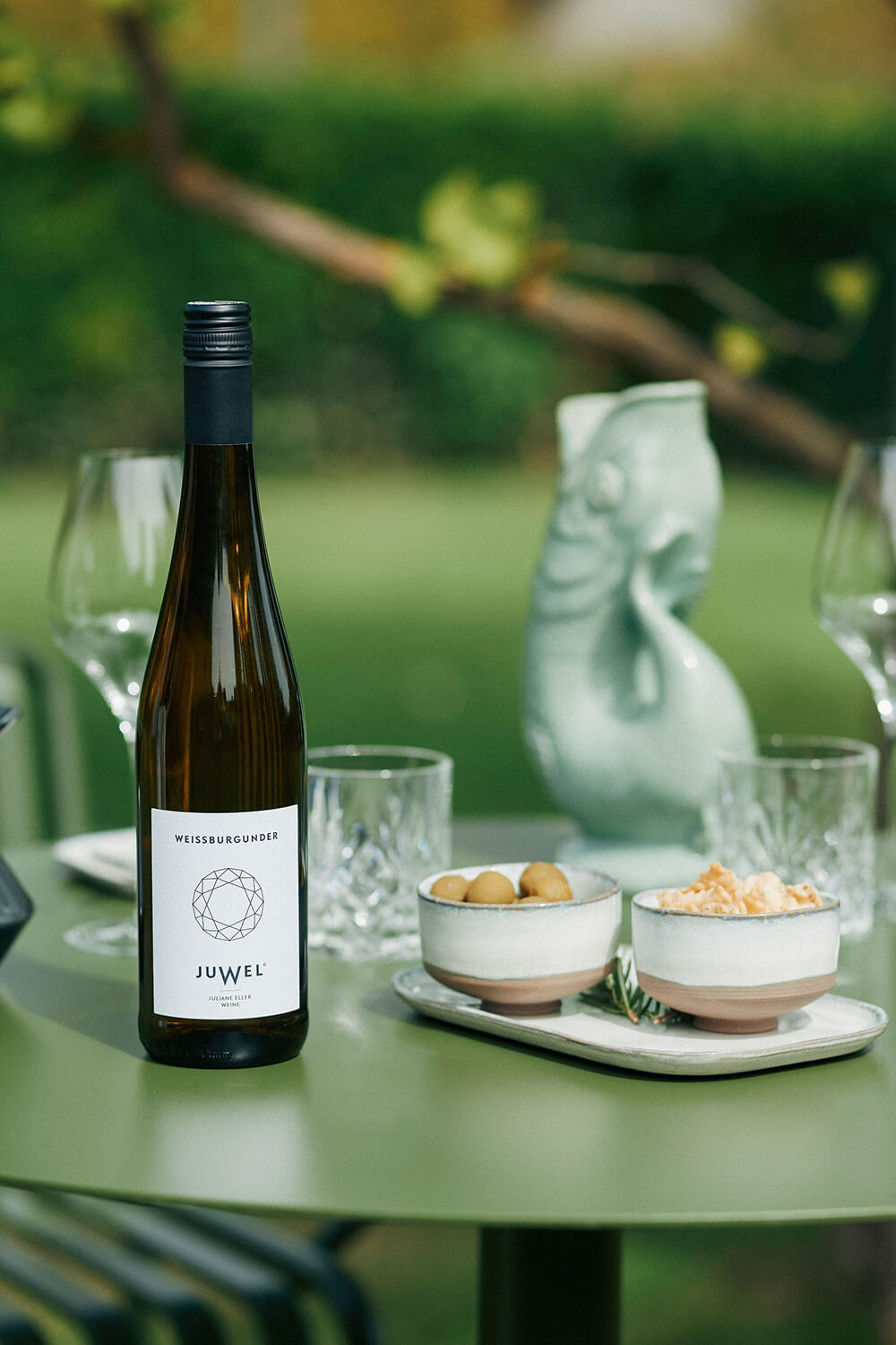 Ein gedeckter Tisch im Garten. Darauf eine Flasche JUWEL Weissburgunder, sowie Schalen mit Oliven und Crackern. Im Hintergrund außerdem Wein- und Wassergläser und ein Wasserkrug