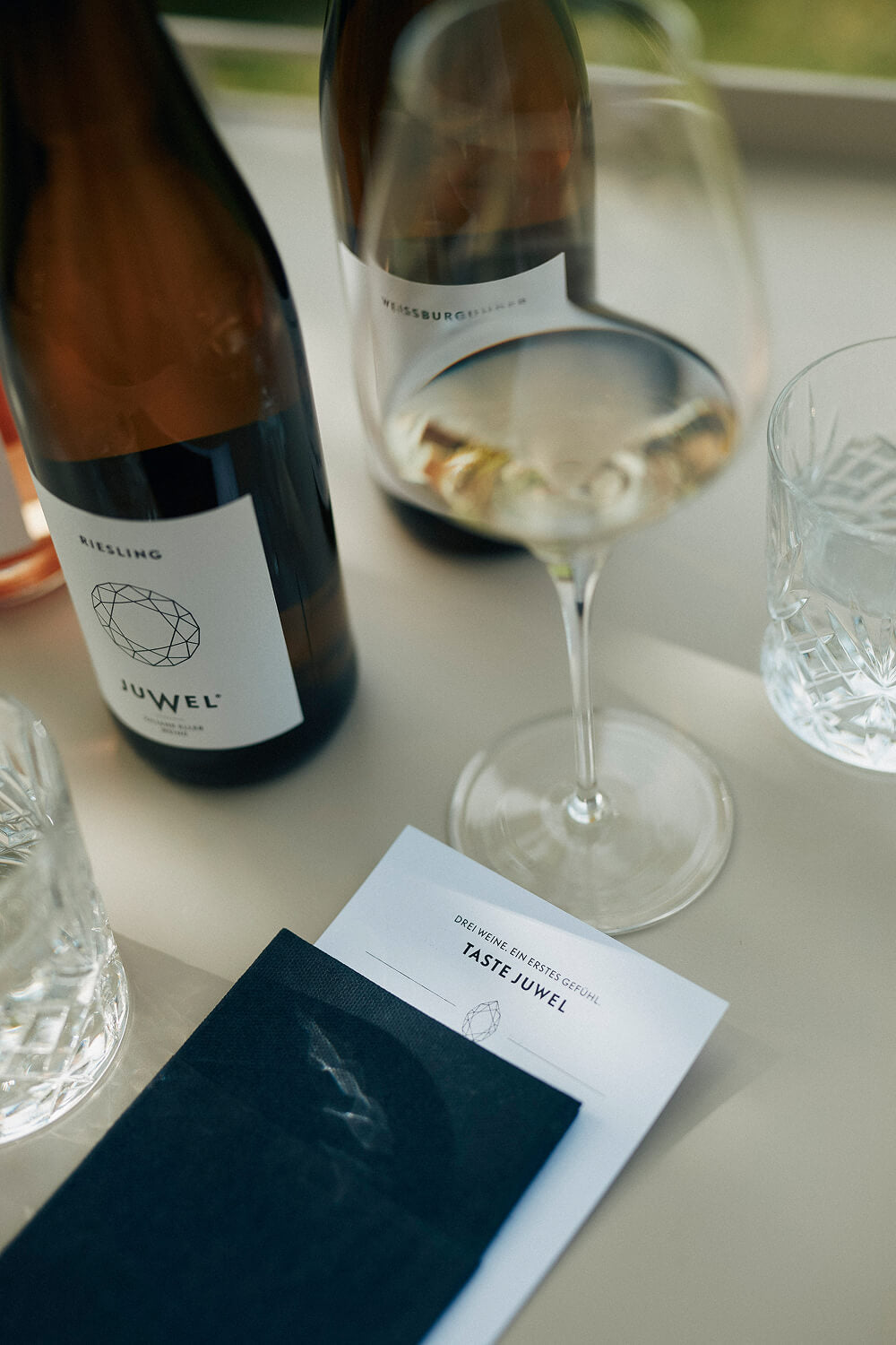 Eine helle Oberfläche mit einer Tastingcard mit der Aufschrift "TASTE JUWEL", die in eine dunkle Serviette gesteckt ist. Darum stehen ein Weißweinglas, Wassergläser und zwei Flaschen JUWEL Wein