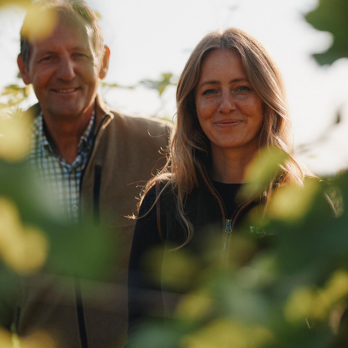 Juliane Eller, Gründerin von JUWEL Weine, steht mit ihrem Vater Thomas im Weinberg und lächelt in die Kamera. Im Vordergrund sind unscharf grüne Blätter zu sehen