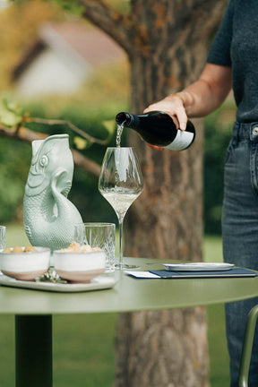 Weinglas im Fokus, das auf einem Tisch im Garten steht. Weißer Wein wird in das Glas geschenkt. Im Vordergrund zwei Schalen mit Oliven und Crackern, im Hintergrund ein Baum
