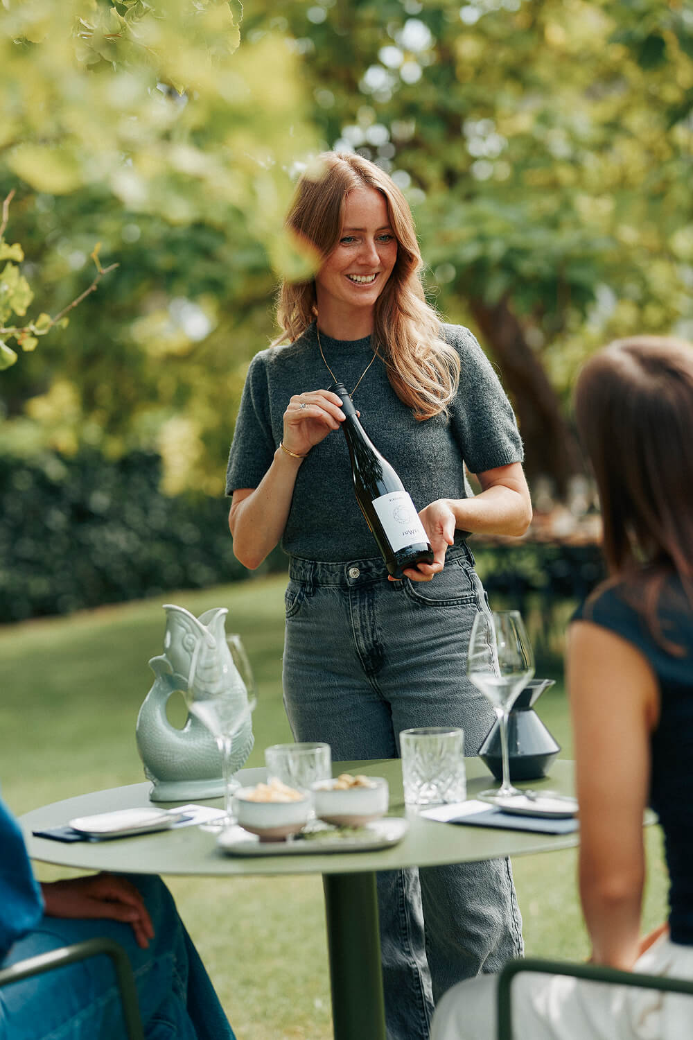 Juliane Eller, Gründerin von JUWEL Weine steht am Tisch im Garten und hält eine Flasche JUWEL Wein in den Händen. Auf dem Zisch stehen Weingläser, kleine Teller und Schalen mit Crackern und Oliven. Juliane spricht mit Personen, die am Tisch sitzen