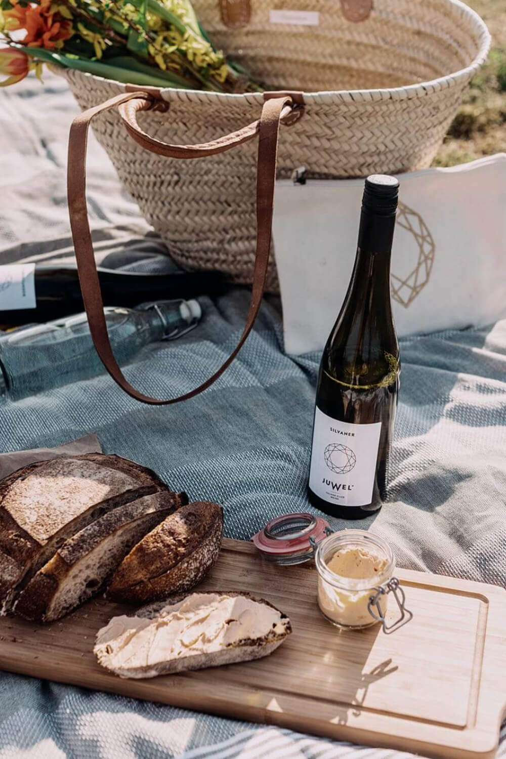 Picknick im Weinberg mit Picknickdecke, Korb, zwei Weinflaschen, ein aufgeschnittenes Brot auf Holzbrett und Aufstrich dabei