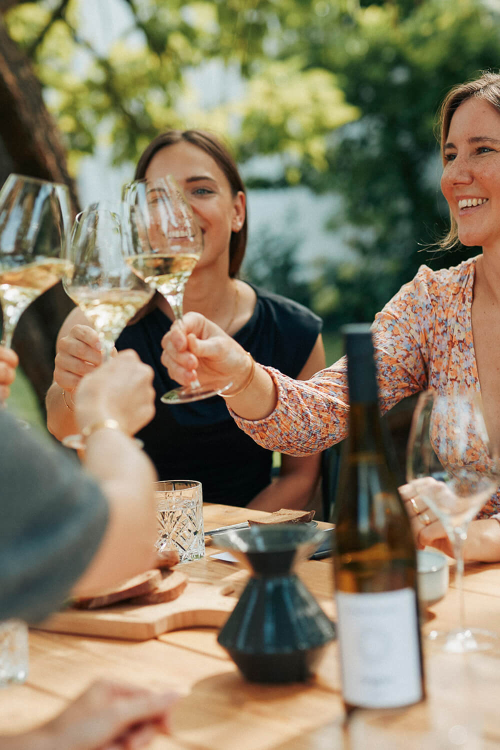 Vier Personen sitzen am gedeckten Tisch im Garten und stoßen mit Weingläsern an. Im Hintergrund verschwommen grüne Bäume, im Vordergrund Weinflasche und aufgeschnittenes Brot