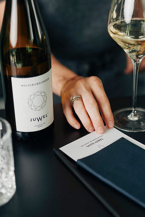 Fokus auf eine dunkle Oberfläche, auf der eine Flasche JUWEL Weissburgunder und ein gefülltes Weißweinglas stehen. Im Fokus eine Tastingcard, die in eine Serviette gesteckt ist, mit der Aufschrift "TASTE JUWEL". Eine Hand berührt die Tastingcard