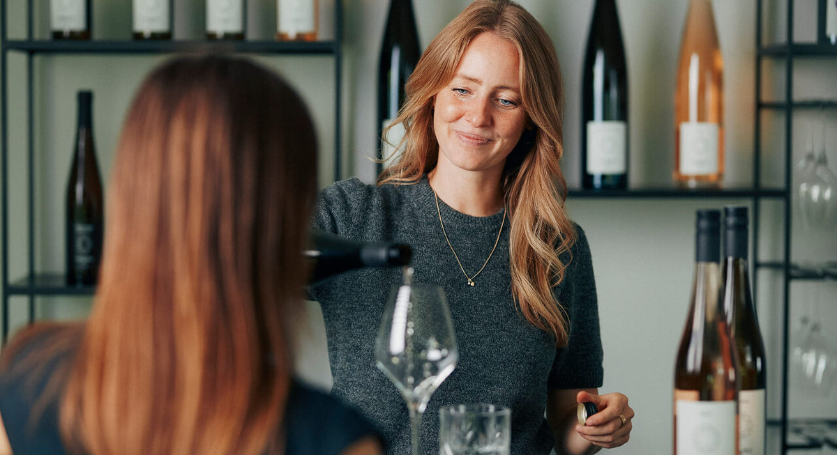 Juliane Eller, Gründerin von JUWEL Weine steht im Verkaufsraum und schenkt einer Frau am Tresen ein Glas Weißwein ein. Im Hintergrund ein modernes Regal mit JUWEL Weinen